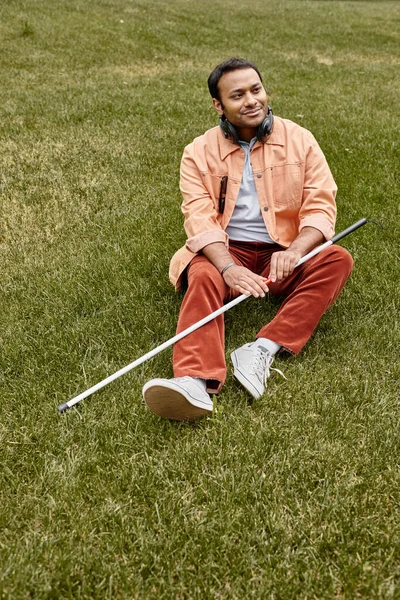 Homem cego indiano alegre em jaqueta laranja sentado na grama com fones de ouvido e bengala — Fotografia de Stock