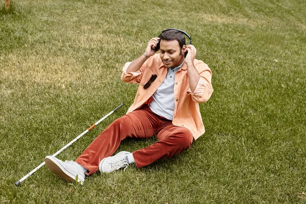 Fröhlicher indischer Blinder in orangefarbener Jacke, der mit Kopfhörern und Gehstock auf dem Gras sitzt — Stockfoto