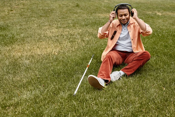 Fröhlicher indischer Blinder in orangefarbener Jacke sitzt mit Kopfhörern und Gehstock im Gras — Stockfoto