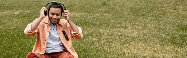 Fröhlicher indischer Blinder in orangefarbener Jacke auf Gras sitzend mit Kopfhörern und Brille, Banner — Stockfoto