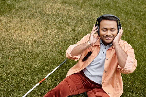 Cheerful indian disabled man in orange jacket sitting on grass with walking stick and enjoying music — Stock Photo