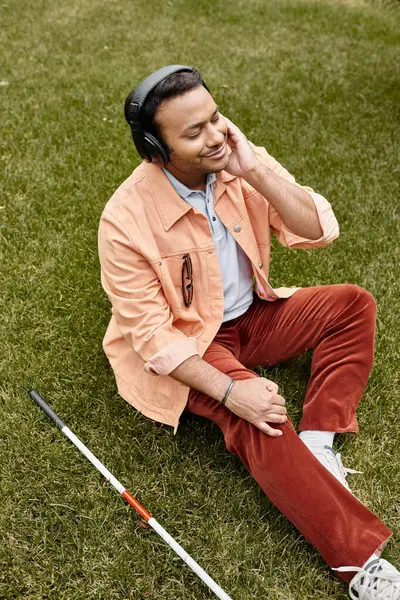 Joyous indian disabled man in orange jacket sitting on grass with walking stick and enjoying music — Stock Photo
