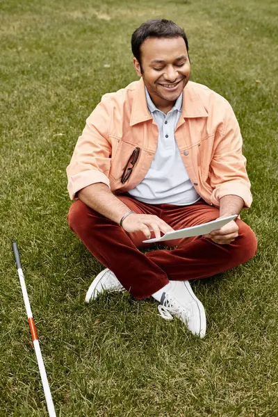 Homem indiano alegre com cegueira em jaqueta laranja sentado na grama e lendo código braille — Fotografia de Stock