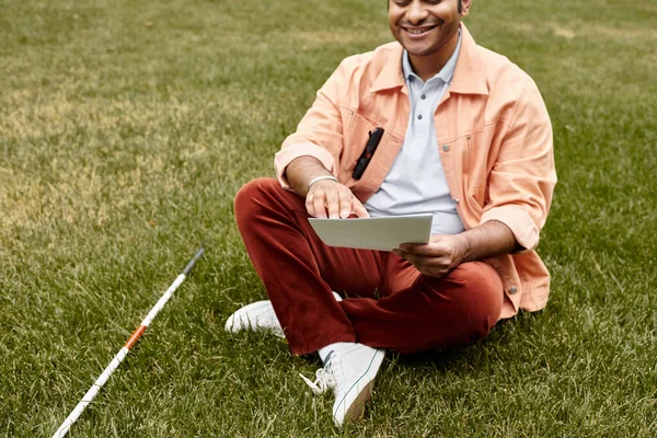 Fröhlicher indischer Mann mit Blindheit in orangefarbener Jacke sitzt auf Gras und liest Braille-Code — Stockfoto