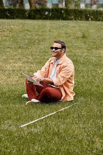 Jolly indian blind man in orange vivid jacket sitting on grass with glasses and reading braille code — Stock Photo