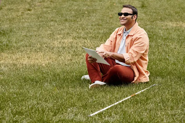 Fröhlicher indischer Blinder in orangefarbener Jacke sitzt mit Brille auf Gras und liest Brailleschrift — Stockfoto