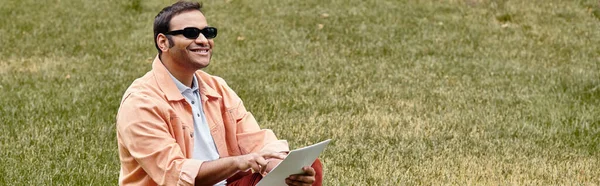 Счастливый индийский слепой в куртке сидит на траве в очках и читает шрифт Брайля, баннер — стоковое фото
