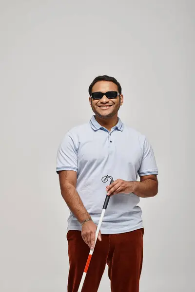 Cheerful indian blind man in blue tee shirt with glasses and walking stick posing on gray backdrop — Stock Photo