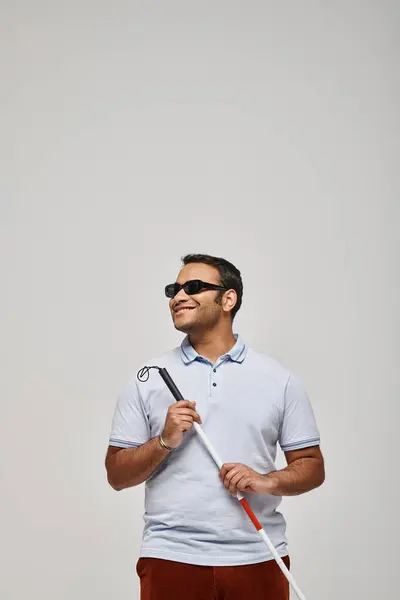 Alegre indio ciego hombre en azul camiseta con gafas y caminando palo posando sobre gris telón de fondo - foto de stock