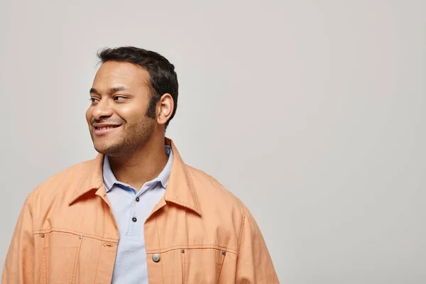 Веселый индийский мужчина в ярко-оранжевой куртке счастливо улыбается и смотрит в сторону на сером фоне — стоковое фото