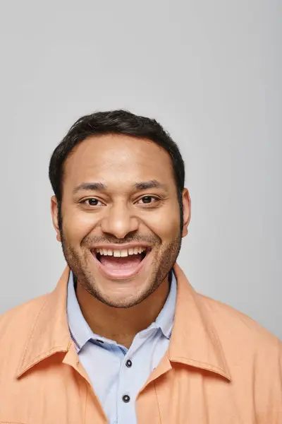 Fröhlich gutaussehender indischer Mann in orangefarbener Jacke lächelt direkt in die Kamera auf grauem Hintergrund — Stockfoto