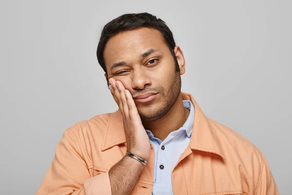 Tired handsome indian man in orange vivid jacket posing with hand on face on gray background — Stock Photo