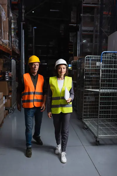 Dos colegas con sombreros duros caminando en un almacén bien iluminado, supervisor y empleado - foto de stock