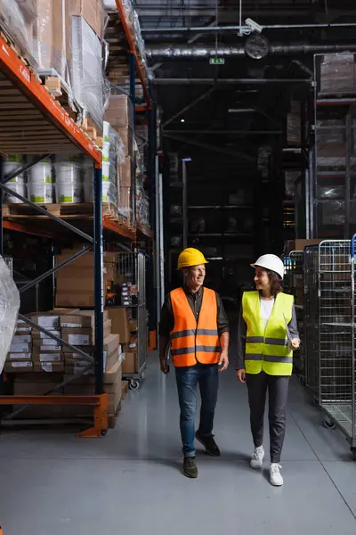 Happy colleagues with hard hats walking in a well-lit warehouse, supervisor and employee — Stock Photo