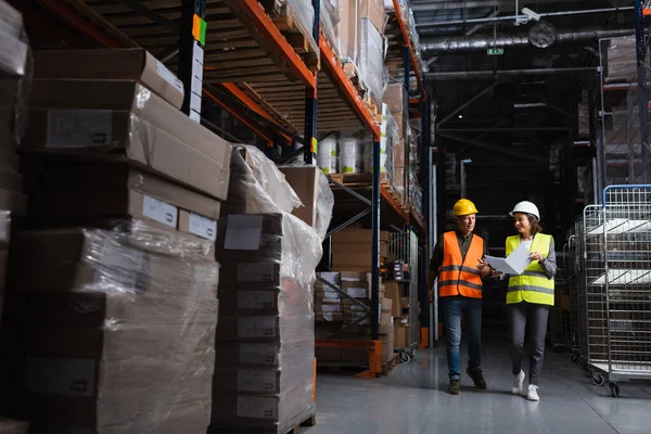 Happy colleagues with hard hats walking in a well-lit warehouse and planning their work, logistic — Stock Photo