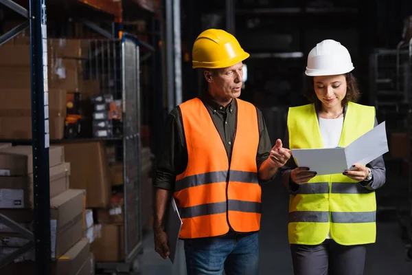Colleagues in safety vests walking in a well-lit warehouse and planning their work, logistic — Stock Photo