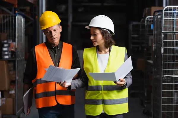 Colleagues in safety vests walking in warehouse and reviewing documents, logistic and cargo — Stock Photo