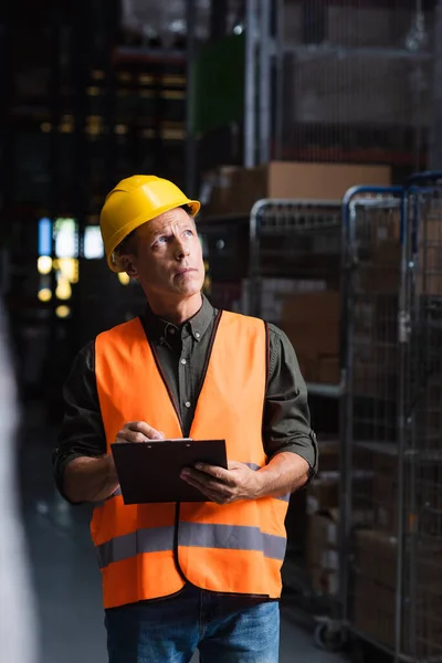 Thoughtful middle aged supervisor in hard hat reviewing paperwork in warehouse, logistics and cargo — Stock Photo