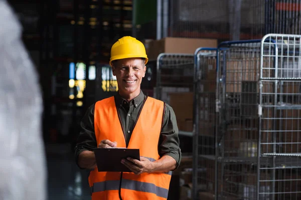 Happy middle aged supervisor in hard hat reviewing paperwork in warehouse, logistics and cargo — Stock Photo