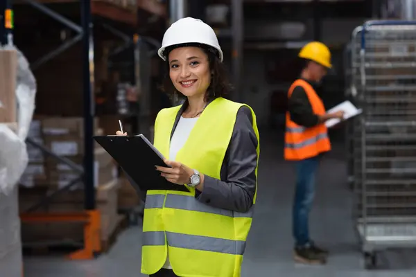 Trabajador de almacén feliz en chaleco de seguridad y escritura de sombrero duro en portapapeles, logística y distribución - foto de stock