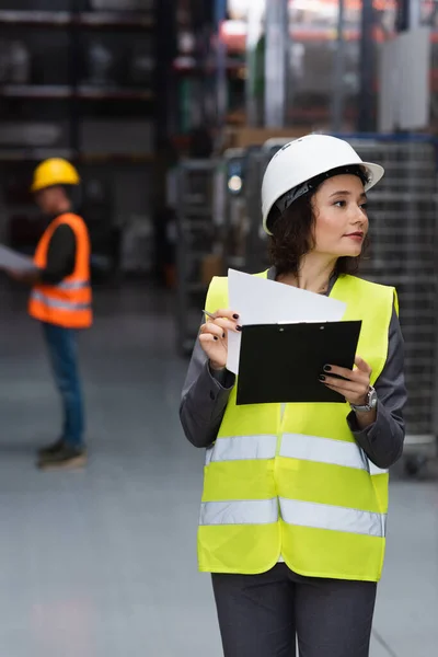 Female warehouse worker in safety vest and hard hat writing on clipboard, logistics and distribution — Stock Photo