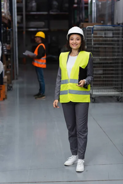 Happy warehouse manager in safety vest and hard hat holding clipboard, logistics and distribution — Stock Photo