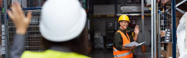 Happy supervisor in hard hat waving at colleague while greeting each other in warehouse, banner — Stock Photo