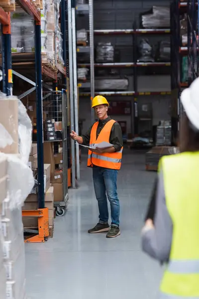 Middle aged supervisor in hard hat with paperwork instructing his female employee in warehouse — Stock Photo