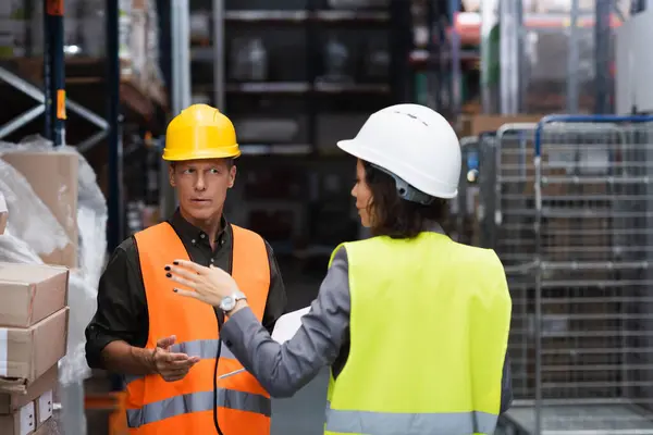 Middle aged supervisor in hard hat having conversation with his female employee in warehouse — Stock Photo