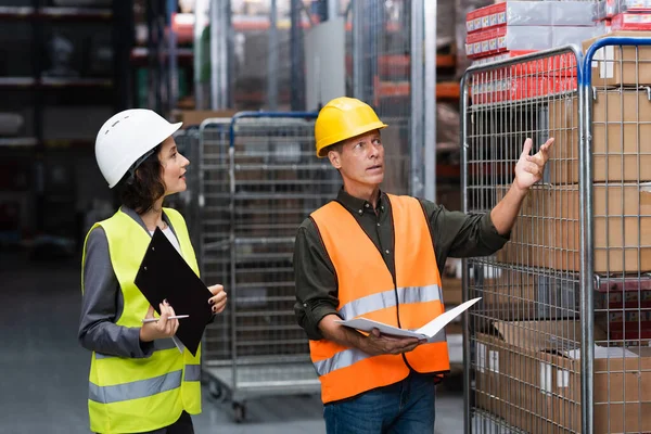 Middle aged supervisor in hard hat with folder instructing his female employee in warehouse — Stock Photo