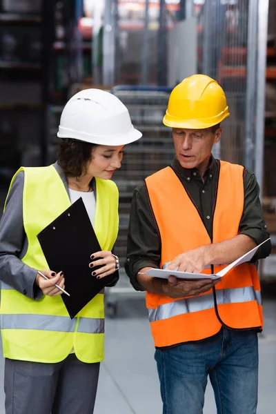 Middle aged supervisor in safety vest showing paperwork to his female employee in warehouse — Stock Photo