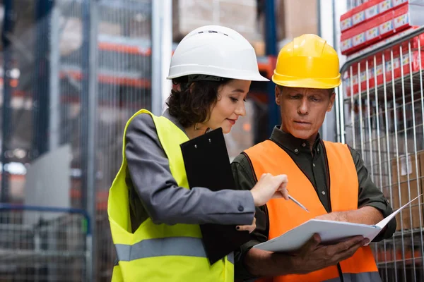 Middle aged warehouse supervisor in safety vest showing paperwork to his female employee with pen — Stock Photo