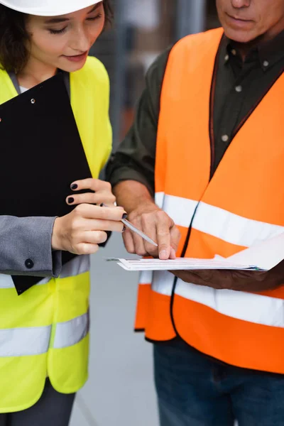 Middle aged warehouse supervisor in safety vest showing paperwork to his female employee with pen — Stock Photo