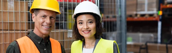 Happy supervisor and employee in hard hats and safety vests looking at camera in warehouse, banner — Stock Photo