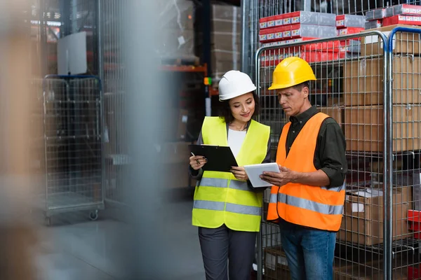 Cheerful supervisor and employee in hard hats standing with clipboard and tablet in warehouse — Stock Photo