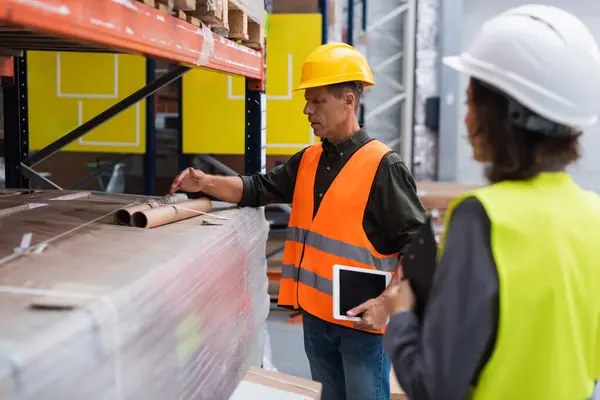 Middle aged supervisor in hard hat holding tablet while explaining work to employee in warehouse — Stock Photo