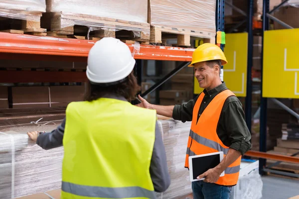 Middle aged supervisor in hard hat holding tablet while explaining work to warehouse employee — Stock Photo
