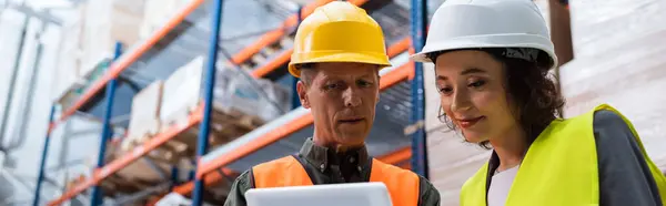 Happy supervisor in hard hat holding tablet while explaining work to employee in warehouse, banner — Stock Photo