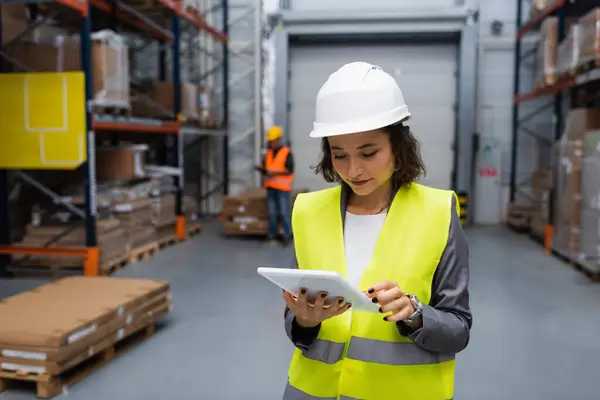 Female warehouse supervisor in hard hat and safety vest focused on digital tablet during work — Stock Photo