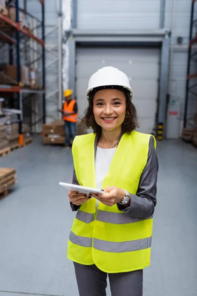 Supervisor armazém feminino feliz em chapéu duro e colete de segurança focado em tablet digital durante o trabalho — Fotografia de Stock