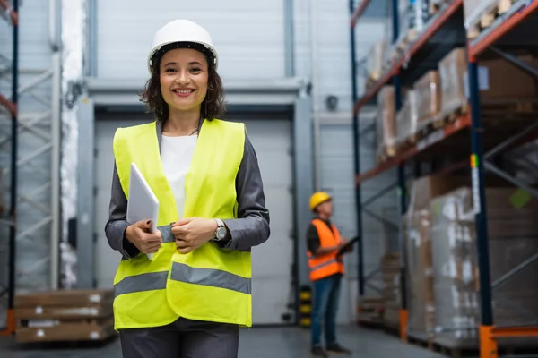 Joyful female warehouse supervisor in hard hat and safety vest focused on digital tablet during work — Stock Photo
