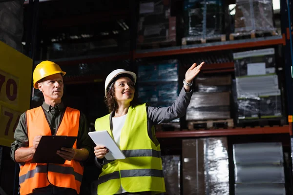 Happy warehouse employee with tablet pointing away near supervisor in hard hat and safety vest — Stock Photo