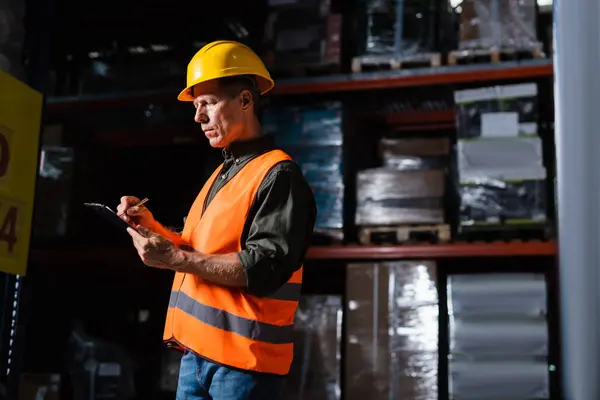 Middle aged warehouse supervisor in hard hat taking notes on clipboard while inspecting cargo — Stock Photo