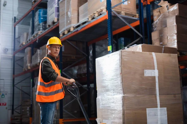 Focused middle aged warehouse worker in hard hat and safety vest transporting pallet with hand truck — Stock Photo