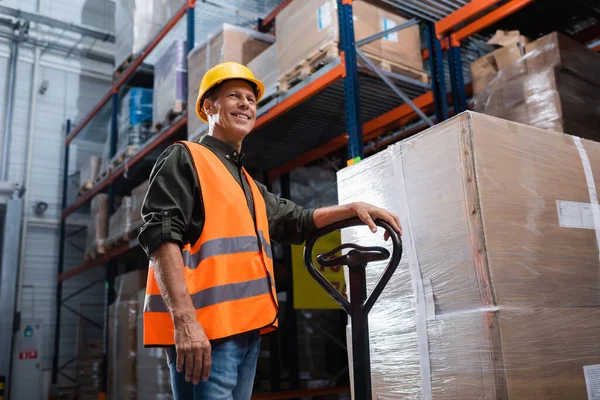 Happy middle aged warehouse worker in hard hat and safety vest transporting pallet with hand truck — Stock Photo