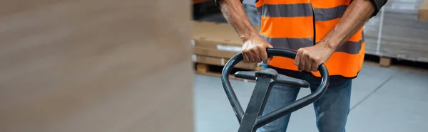 Cropped banner of warehouse worker in hard hat and safety vest transporting pallet with hand truck — Stock Photo