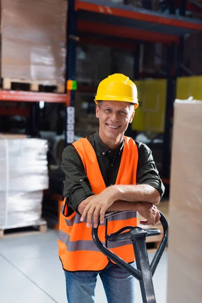 Travailleur d'entrepôt joyeux en casque dur et gilet de sécurité transportant palette avec camion à main, sourire — Photo de stock