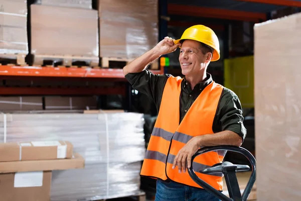 Щасливий складський робітник у твердій шапці та жилеті, що перевозить піддон з ручною вантажівкою, посмішка — стокове фото