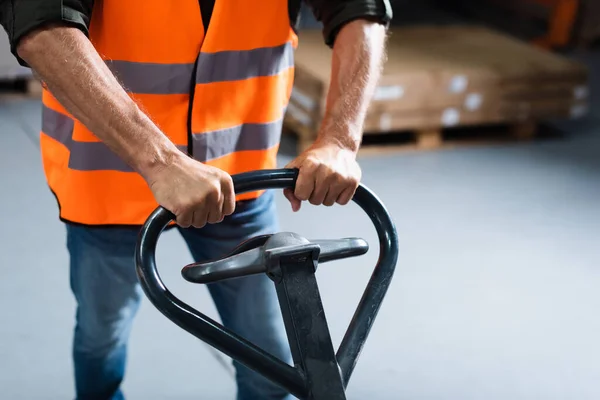 Cropped shot of warehouse worker in jeans and safety vest transporting pallet with hand truck — Stock Photo