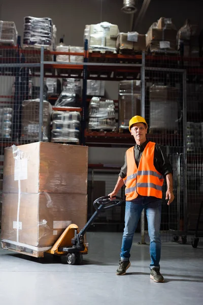 Travailleur d'entrepôt d'âge moyen en casque dur et gilet de sécurité transportant palette avec camion à main, sourire — Photo de stock